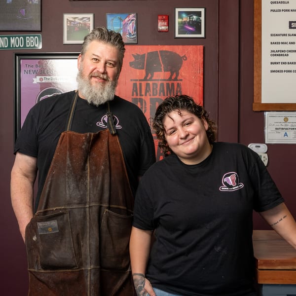 Oink And Moo BBQ owners, a man and woman, smiling in front of a rustic wall with restaurant decor.