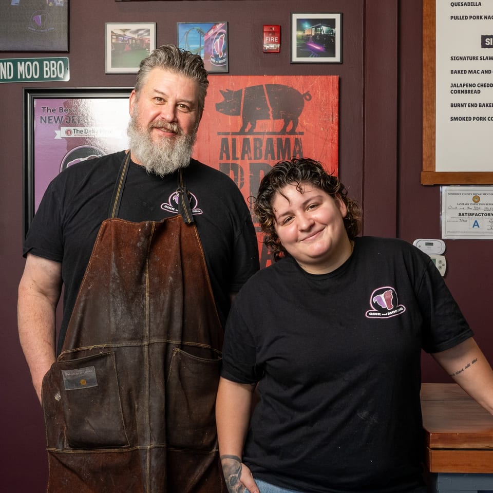 Oink And Moo BBQ owners, a man and woman, smiling in front of a rustic wall with restaurant decor.