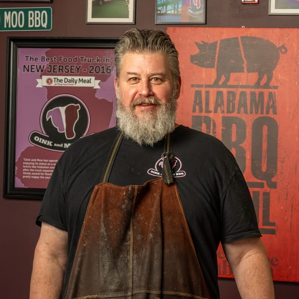 A man with a gray beard and black Oink and Moo BBQ shirt smiles, wearing a brown apron.