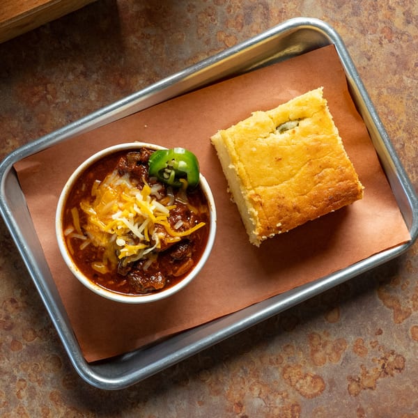 A bowl of Oink And Moo BBQ chili with cheese and jalapeños, alongside a piece of cornbread.